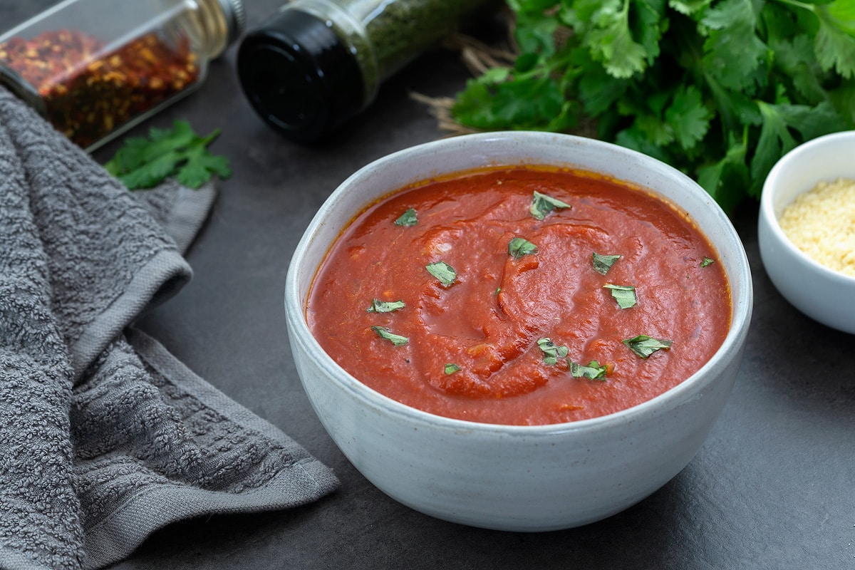 Marinara Sauce in a white bowl placed on a gray table with few ingredients around